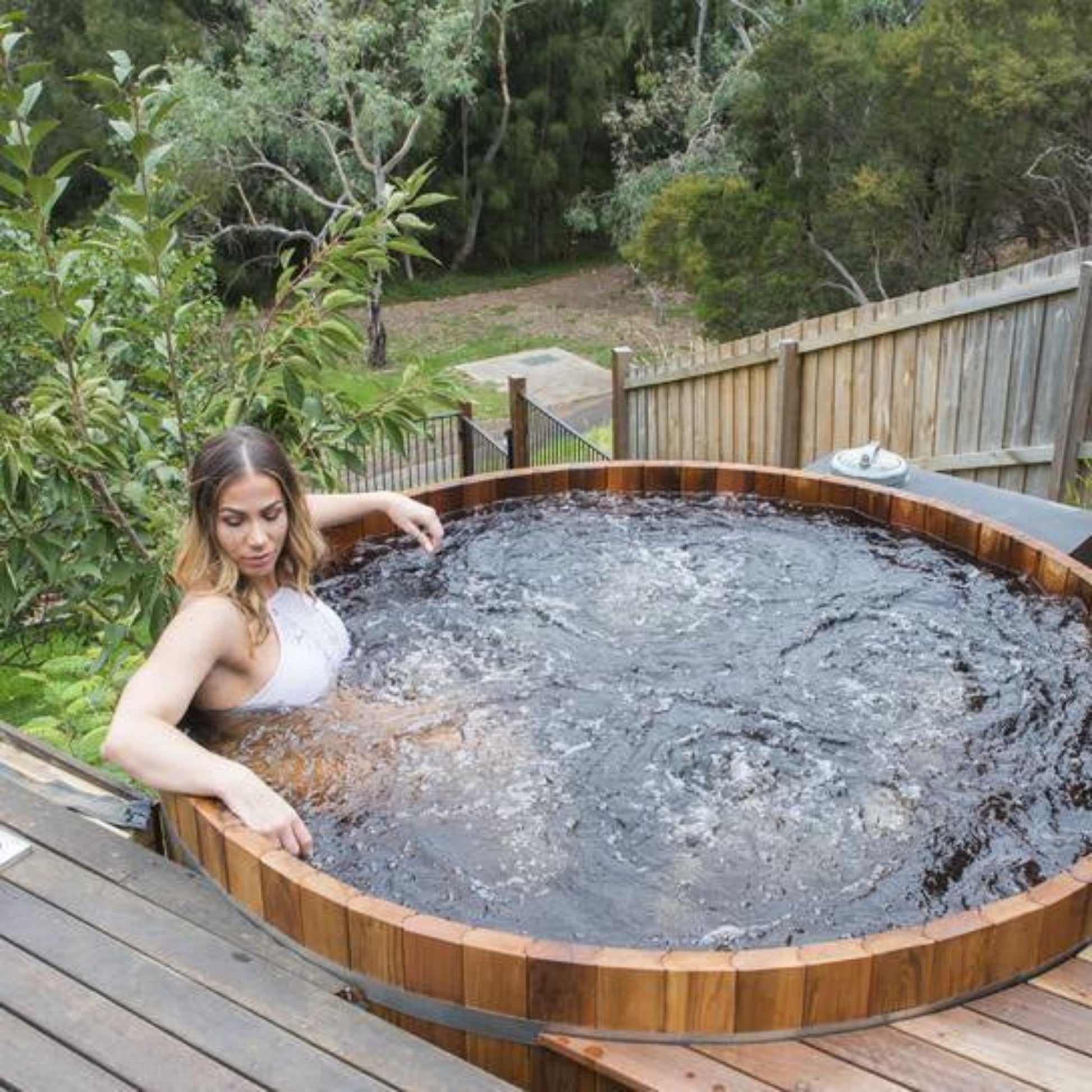 Woman relaxing in a wooden hot tub outdoors with greenery in the background