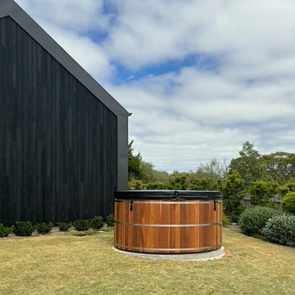 Wooden hot tub in a garden with a black building and trees in the background